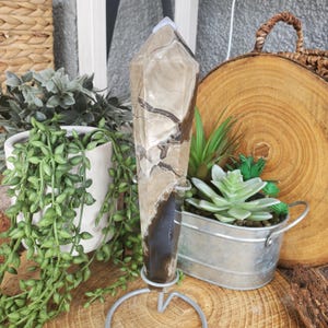May include: A tall, polished crystal point with a light brown and dark brown marbled pattern. The crystal is mounted on a silver metal stand. The crystal is a decorative item.