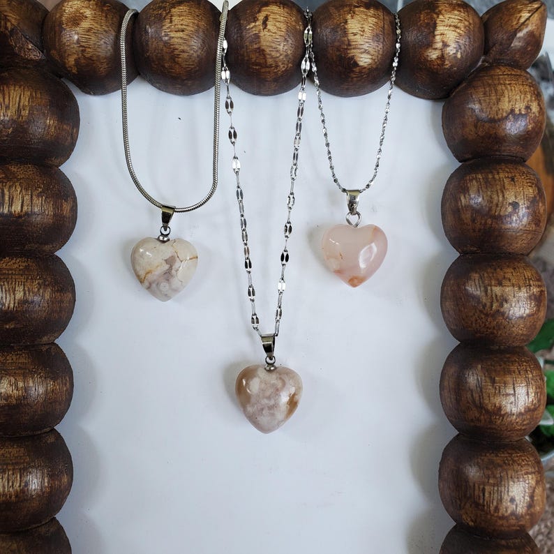 Op de afbeelding: Drie kettingen met hartvormige hangers. De hangers zijn gemaakt van een lichtgekleurde steen met verschillende patronen. De kettingen zijn zilverkleurig. De kettingen worden getoond op een houten frame met een donkerbruine afwerking.