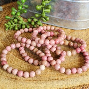 May include: A set of six pink gemstone bracelets made of round beads. The bracelets are stacked on top of each other on a wooden surface.