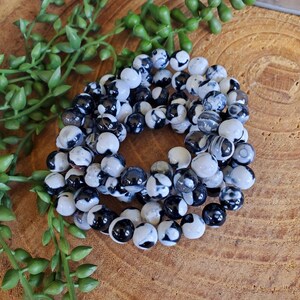 May include: A collection of black and white beaded bracelets. The beads are round and have a marbled appearance. The bracelets are arranged in a circular pattern on a wooden surface, with green leafy sprigs in the background.