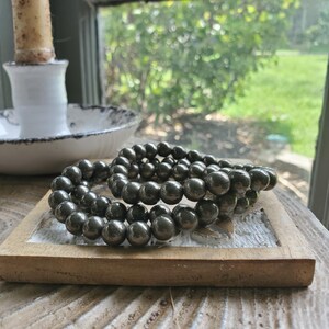 May include: Two dark gray beaded bracelets rest on a small, light brown wooden tray. The bracelets are made of round, polished beads. A white candle holder and a window with a blurred green background are visible in the background.