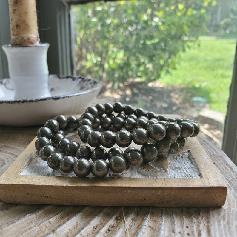 May include: Two dark gray beaded bracelets rest on a small, light brown wooden tray. The bracelets are made of round, polished beads. A white candle holder and a window with a blurred green background are visible in the background.