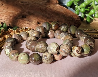 Puede incluir: Un brazalete de cuentas redondas de jaspe con patrones en tonos marrones, verdes y crema. El brazalete se exhibe sobre una superficie rosa, con una pieza de madera y vegetación en el fondo.