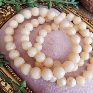 May include: Three strands of cream-colored beaded bracelets. The beads are round and have a smooth, matte finish. The bracelets are arranged on a pink surface, with a gold-colored decorative frame in the background. The beads appear to be approximately 0.8 cm in diameter.