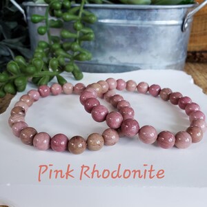 May include: Two pink rhodonite beaded bracelets. The bracelets are made of round, smooth beads. The text "Pink Rhodonite" is written below the bracelets.