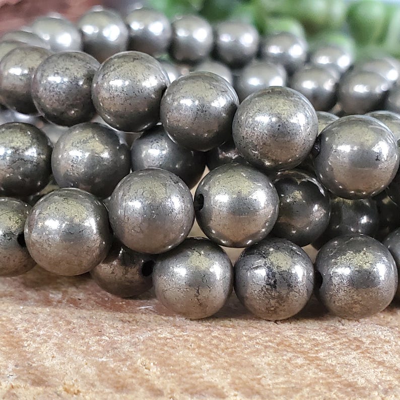 May include: Close-up of a strand of pyrite beads. The beads are spherical, with a metallic silver-gray color and a slightly textured surface. The beads are strung together, suggesting they are for jewelry making or crafting.