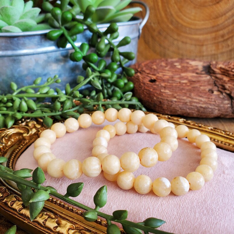 May include: Two cream-colored beaded bracelets rest on a pink surface within a gold-colored frame. The bracelets are composed of round beads. Green plants and a small metal bucket are in the background.