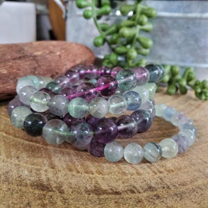 May include: Several bracelets made of round, polished fluorite beads in shades of purple, green, and clear. The beads are strung on elastic, creating a stackable accessory. The bracelets are displayed on a wooden surface.