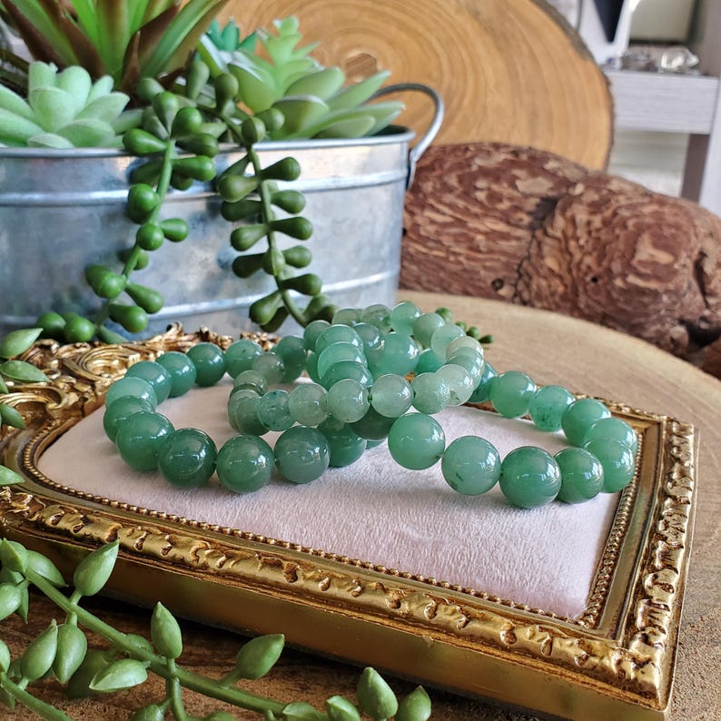 May include: Three jade green beaded bracelets rest on a gold-framed, pink velvet surface. The bracelets are composed of smooth, round beads. A galvanized metal bucket with green succulents and trailing plants is in the background.