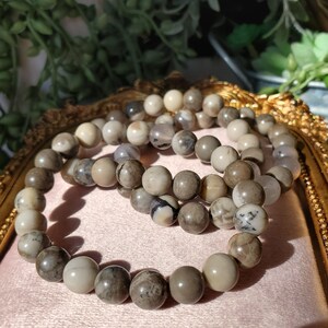 May include: Three beaded bracelets with neutral-toned, round beads. The beads vary in color, including cream, gray, and brown, with some displaying unique patterns. The bracelets are arranged on a pink surface.