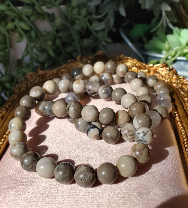 May include: Three beaded bracelets with neutral-toned, round beads. The beads vary in color, including cream, gray, and brown, with some displaying unique patterns. The bracelets are arranged on a pink surface.