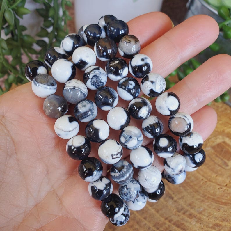 May include: A collection of round beads in shades of black, white, and gray. The beads have a marbled appearance, with swirling patterns and variations in color. The beads are smooth and polished, and appear to be made of stone or a similar material.