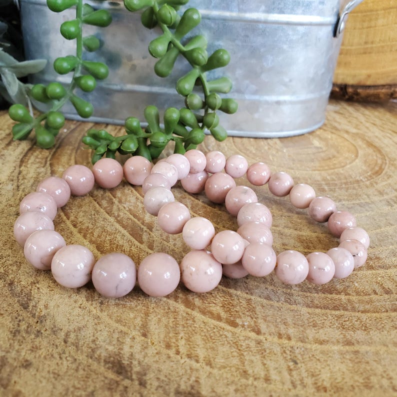 Puede incluir: Dos pulseras de piedras preciosas rosas hechas con cuentas redondas. Las pulseras est&aacute;n apiladas una encima de la otra.