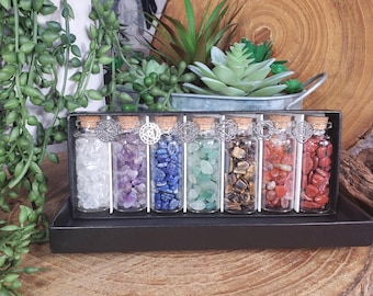 May include: A collection of six glass bottles filled with different coloured gemstones, such as amethyst and lapis lazuli. Each bottle has a cork stopper and silver charms. The bottles are displayed in a black box, presented on a wooden surface.