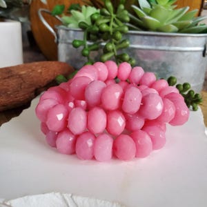 May include: A stack of three pink beaded bracelets. The beads are faceted, giving them a sparkling appearance. The bracelets are displayed on a white surface, with a blurred background of decorative items.