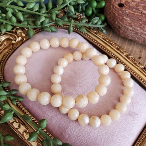 May include: Two cream-colored beaded bracelets rest on a pink surface within a gold-colored ornate frame. The bracelets are made of round, smooth beads. Green foliage and a decorative object are in the background.