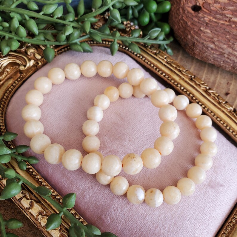 May include: Two cream-colored beaded bracelets rest on a pink surface within a gold-colored ornate frame. The bracelets are made of round, smooth beads. Green foliage and a decorative object are in the background.