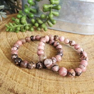 Puede incluir: Dos pulseras de piedras preciosas rosas y negras con cuentas redondas. Las pulseras est&aacute;n hechas de piedras naturales y est&aacute;n apiladas una encima de la otra.