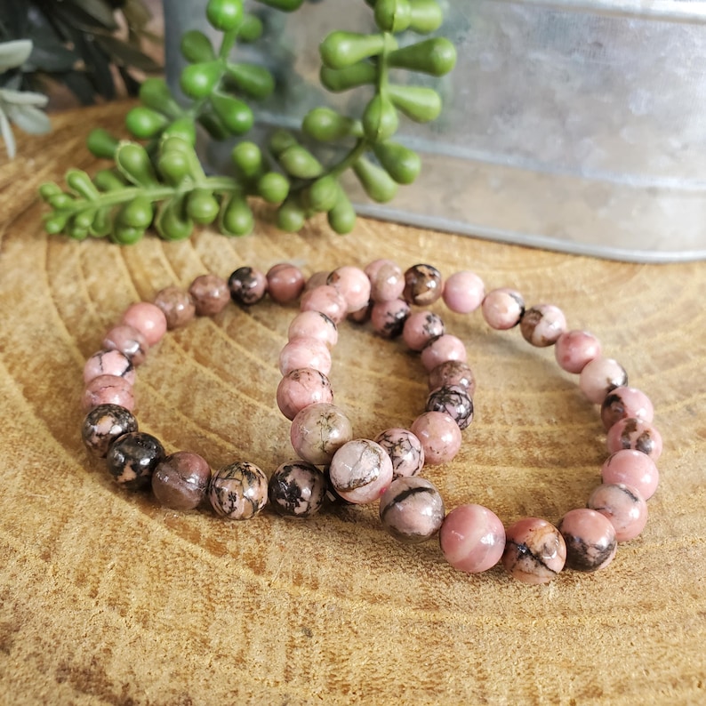 Puede incluir: Dos pulseras de piedras preciosas rosas y negras con cuentas redondas. Las pulseras est&aacute;n hechas de piedras naturales y est&aacute;n apiladas una encima de la otra.