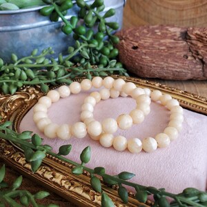May include: Two cream-colored beaded bracelets rest on a pink surface within a gold-colored frame. The bracelets are made of round beads. Green leafy plants and a small metal bucket are in the background.