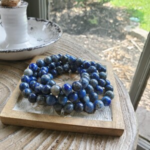 May include: A collection of blue and gray beaded bracelets arranged on a small, rustic wooden tray. The beads are round and have a marbled appearance. The tray sits on a wooden surface, with a white ceramic container in the background.