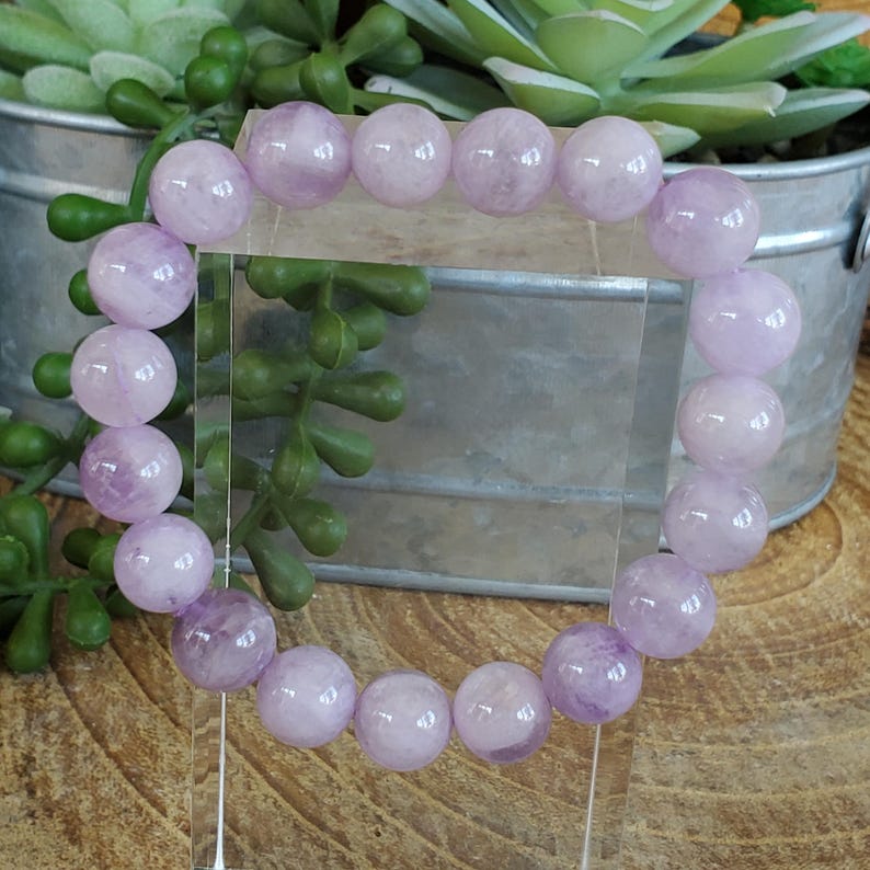 K&ouml;nnte beinhalten: Ein helllila Perlenarmband mit runden, polierten Steinen. Das Armband wird auf einem transparenten St&auml;nder pr&auml;sentiert, mit einem Metalleimer und gr&uuml;nen Pflanzen im Hintergrund. Die Perlen haben eine durchscheinende Qualit&auml;t.