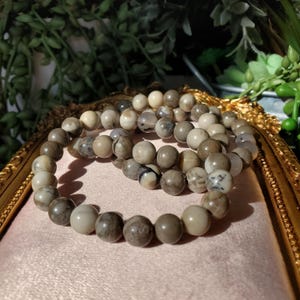 May include: Three beaded bracelets with neutral-toned, round beads. The beads have variations in color, including cream, gray, and brown. The bracelets are arranged on a pink surface within a gold-framed decorative tray, with greenery in the background.
