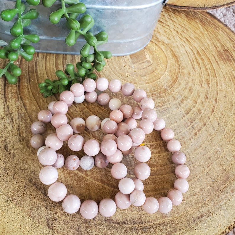 Puede incluir: Tres pulseras de piedras preciosas rosas hechas de cuentas redondas. Las pulseras est&aacute;n apiladas una encima de la otra sobre una superficie de madera.