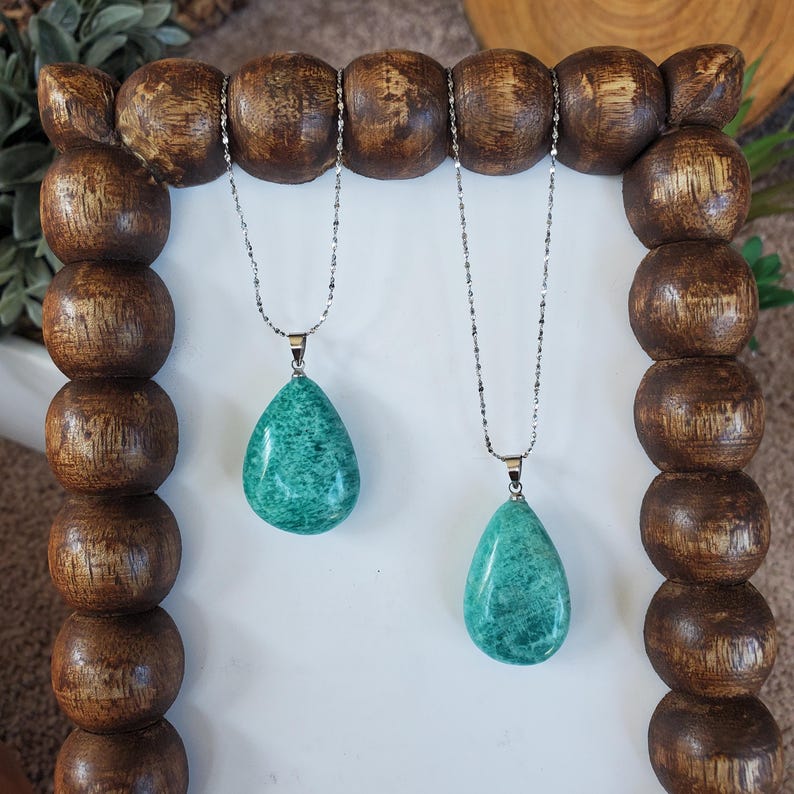 May include: Two necklaces with teal teardrop pendants hang from a wooden display. The pendants have a textured appearance and are suspended from delicate silver chains. The wooden display has a light brown finish and a unique, rounded design.