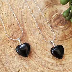 May include: Two silver chain necklaces with black heart-shaped pendants. The necklaces are on a wooden surface.