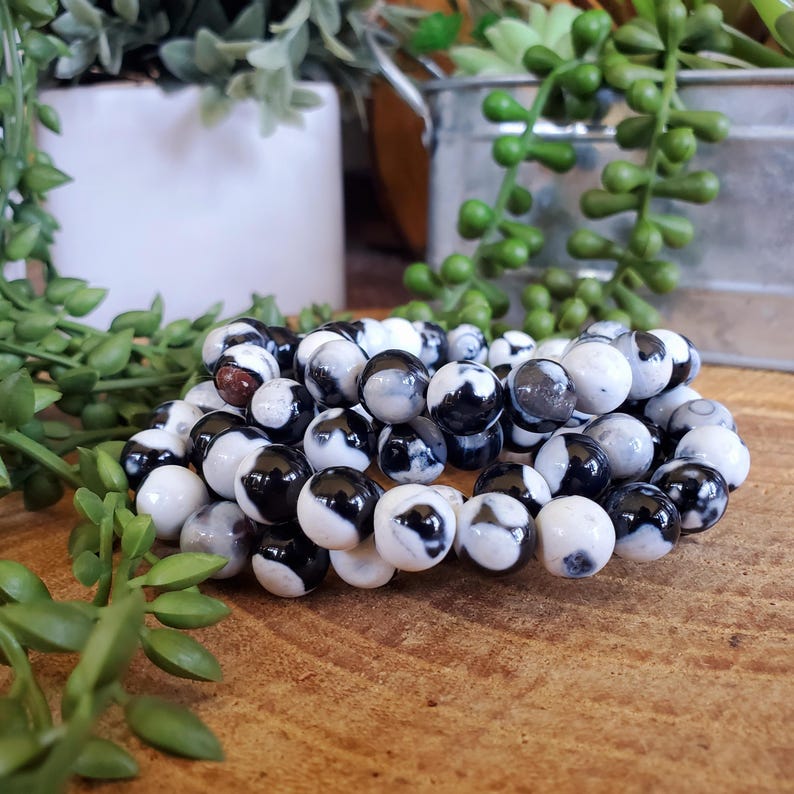May include: A stack of round bracelets made of black and white patterned beads. The beads have a marbled appearance, with swirls of black and white. The bracelets are resting on a wooden surface, with greenery in the background.