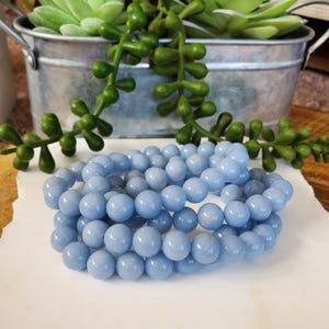 May include: A collection of light blue, round beads, likely for jewelry making, are arranged on a white surface. The beads are smooth and evenly colored. Green succulent plants in a metal bucket are in the background.