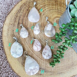 May include: A collection of eight teardrop-shaped gemstone pendants with silver-colored bails. The pendants feature various patterns of white and pink hues. The pendants are displayed on a wooden surface, with a small metal bucket and greenery in the background.