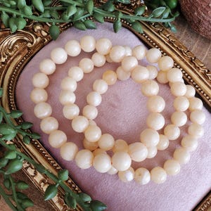 May include: Three cream-colored beaded bracelets are arranged on a pink surface within a gold-colored ornate frame. The bracelets are made of round, smooth beads. Green foliage accents the frame.