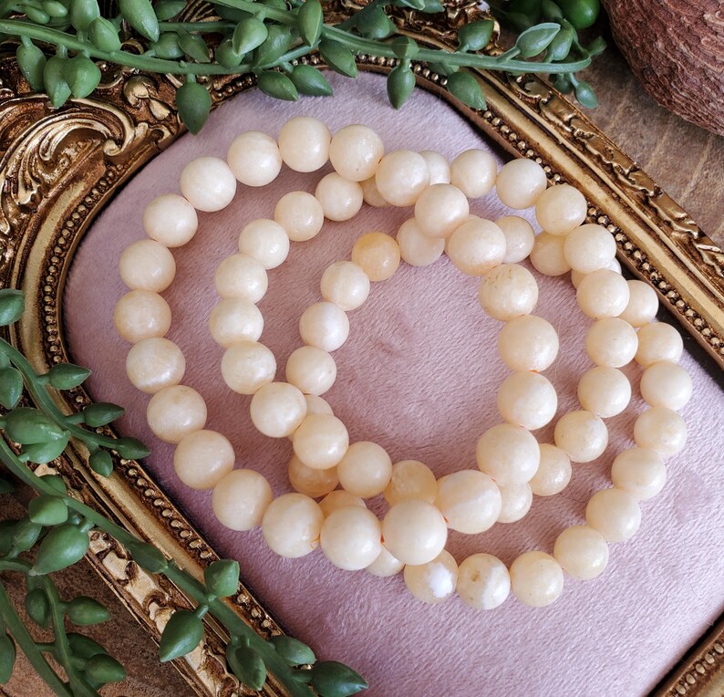 May include: Three cream-colored beaded bracelets are arranged on a pink surface within a gold-colored ornate frame. The bracelets are made of round, smooth beads. Green foliage accents the frame.
