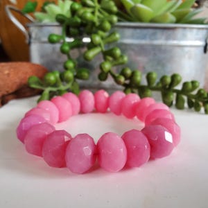 May include: A pink beaded bracelet with faceted, oval-shaped beads. The bracelet is a gradient of pink hues, from light to darker shades. The beads are strung together, forming a complete circle. The bracelet is displayed on a white surface.