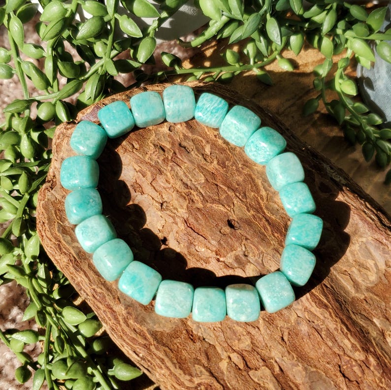 Puede incluir: Un brazalete de Amazonita turquesa compuesto por cuentas cuadradas. El brazalete se exhibe sobre un trozo de madera, con follaje verde en el fondo. Las cuentas miden aproximadamente 1,3 cm.