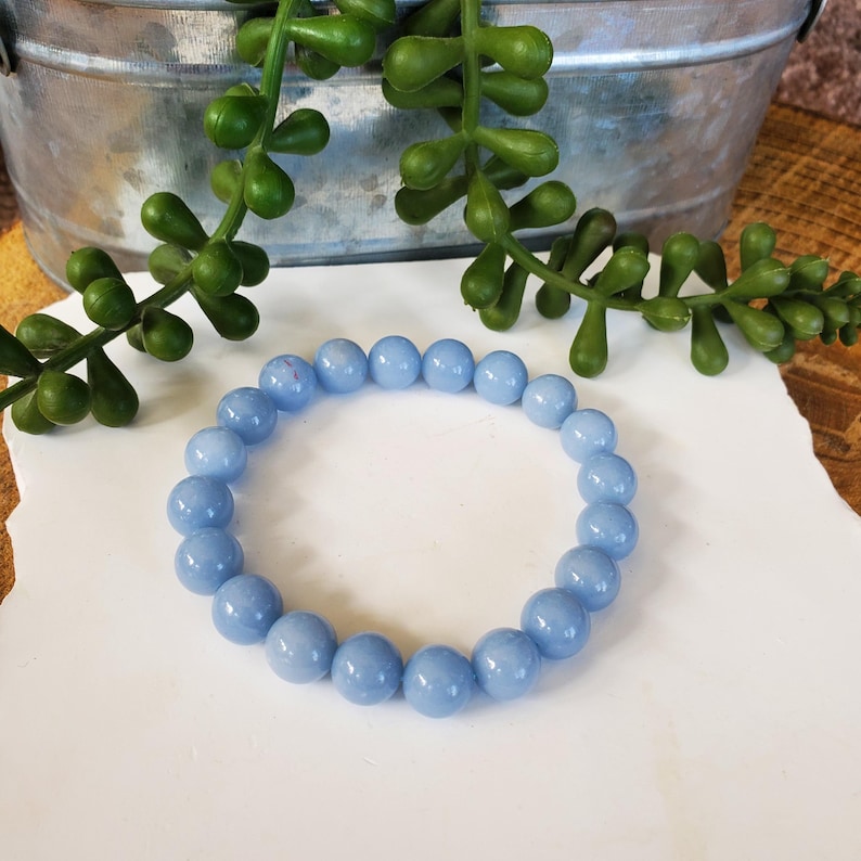 May include: A light blue beaded bracelet. The bracelet is made of round, polished beads. The beads are a soft, pastel blue color. The bracelet is displayed on a white surface with green plant accents.