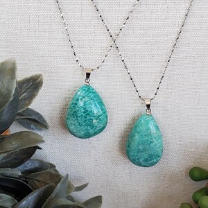 May include: Two necklaces featuring turquoise teardrop pendants on delicate silver chains. The pendants have a marbled appearance. The background is a neutral off-white fabric. Green plants are visible in the foreground.