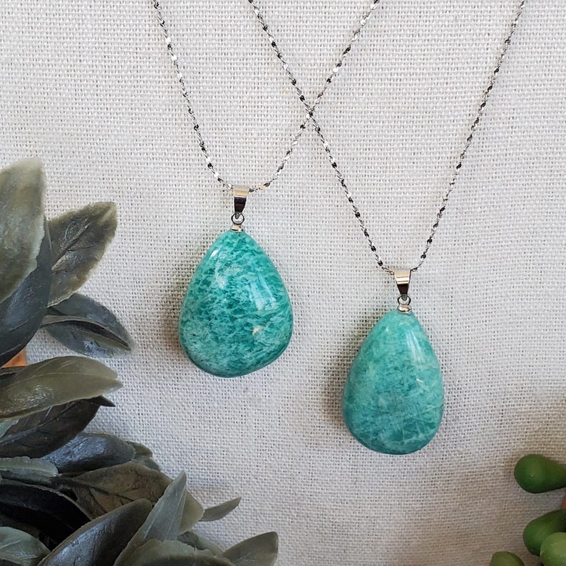 May include: Two necklaces featuring turquoise teardrop pendants on delicate silver chains. The pendants have a marbled appearance. The background is a neutral off-white fabric. Green plants are visible in the foreground.
