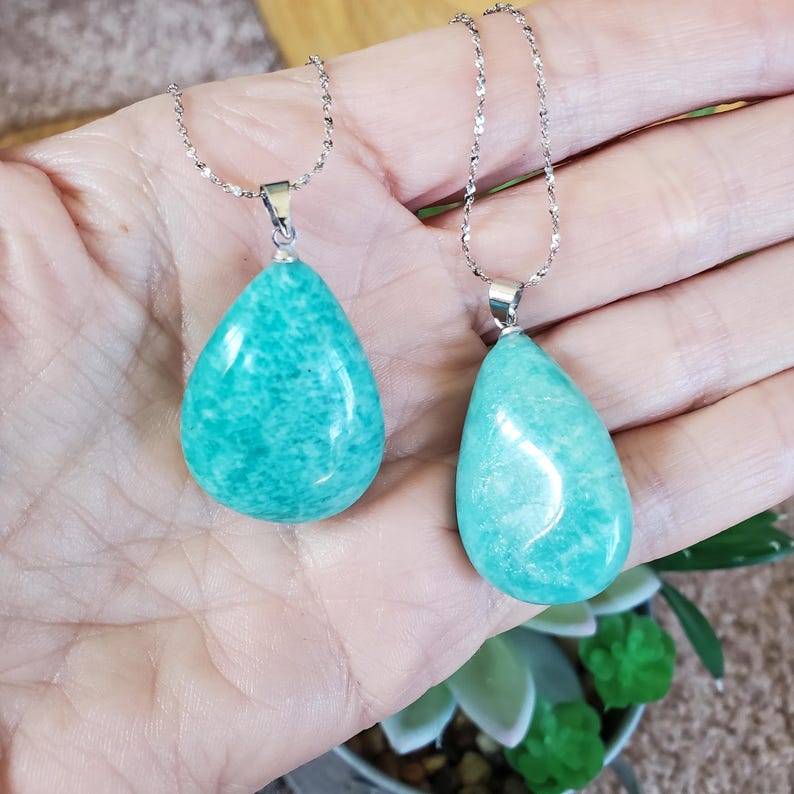 May include: Two teal teardrop pendants on delicate silver chains. The pendants have a smooth, polished surface. The jewellery is displayed against a neutral background. The pendants are a light teal colour.
