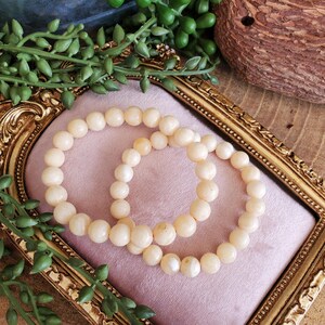 May include: Two cream-colored beaded bracelets rest on a pink velvet surface within a gold-framed decorative tray. The bracelets are composed of round, smooth beads. Green leafy vines and a decorative wooden object are in the background.