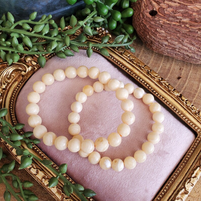 May include: Two cream-colored beaded bracelets rest on a pink velvet surface within a gold-framed decorative tray. The bracelets are composed of round, smooth beads. Green leafy vines and a decorative wooden object are in the background.