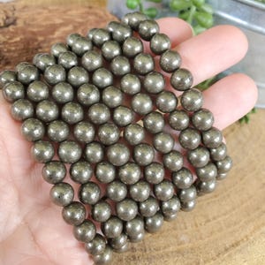 May include: Strand of pyrite beads. The beads are spherical, with a metallic, silvery-gold color. The beads are held in a hand, and the background is a wooden surface.