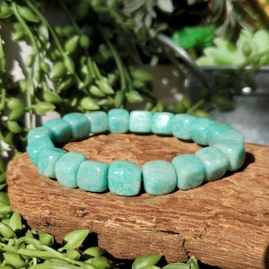 Puede incluir: Un brazalete de piedras preciosas turquesas compuesto por cuentas cuadradas. El brazalete se exhibe sobre un trozo de madera desgastada, con follaje verde en el fondo. El brazalete es de color azul verdoso claro.