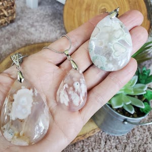 May include: Three agate gemstone pendants with silver-colored chains. The pendants are teardrop-shaped, showcasing a mix of colors including white, pink, and gray. The gemstones have unique patterns and textures, and are displayed on a wooden surface.
