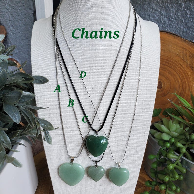 Op de afbeelding: Vier kettingen met hartvormige hangers worden tentoongesteld op een mannequin. De hangers zijn in verschillende groentinten. De kettingen zijn zilver en zwart. Het woord "Chains" staat in het groen boven de kettingen.