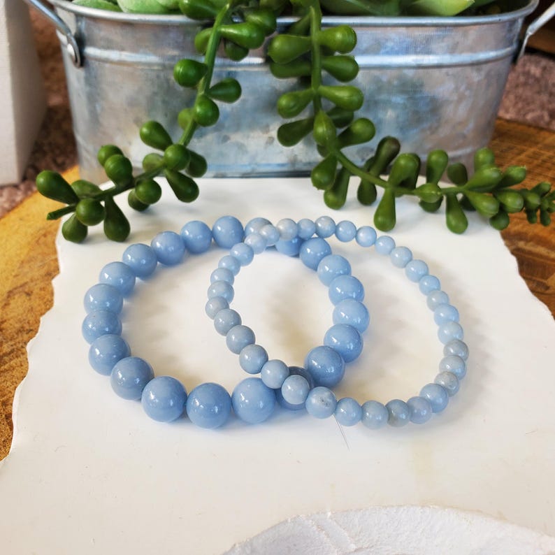 May include: Two light blue beaded bracelets. One bracelet has larger beads, while the other has smaller beads. The bracelets are displayed on a white surface, with a decorative metal container and green plant in the background.