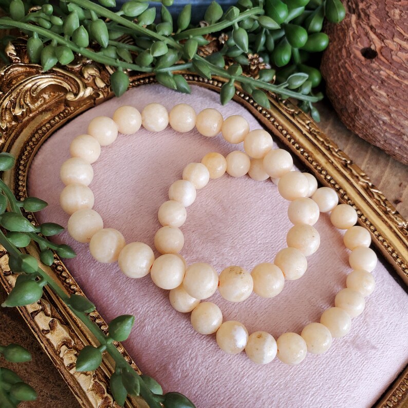 May include: Two beaded bracelets with cream-colored, round beads. The bracelets are displayed on a pink surface within a gold-colored, ornate frame. Greenery is visible in the background, adding a natural touch to the presentation.