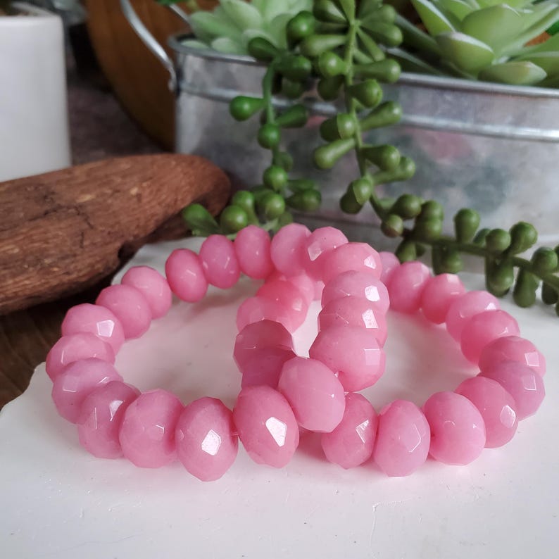 May include: Two pink faceted bead bracelets. The beads are a light pink color and have a faceted cut. The bracelets are displayed on a white surface, with greenery and a metal container in the background.